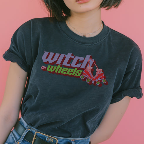 Gray t-shirt with "Witch on Wheels" text in purple and green, featuring a red roller skate graphic. The vintage-style design has a slightly faded look on a relaxed-fit unisex tee.